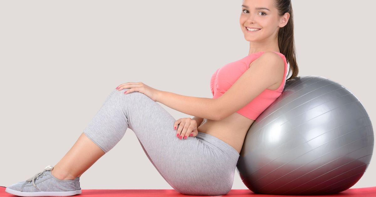 Girl leaning on a Pilates ball, sliming at the camera.