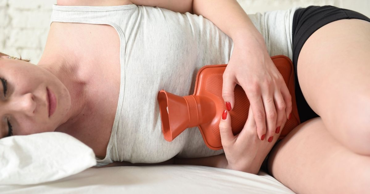Image of a young woman alleviating period cramps by using a hot water bottle on her stomach while lying down.