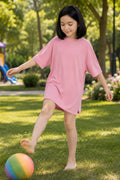 A girl wearing an oversized light pink t-shirt dress and kicking a colorful rainbow ball on the green grass.