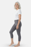 A woman wearing gray capri leggings and a white top against a light gray studio background.