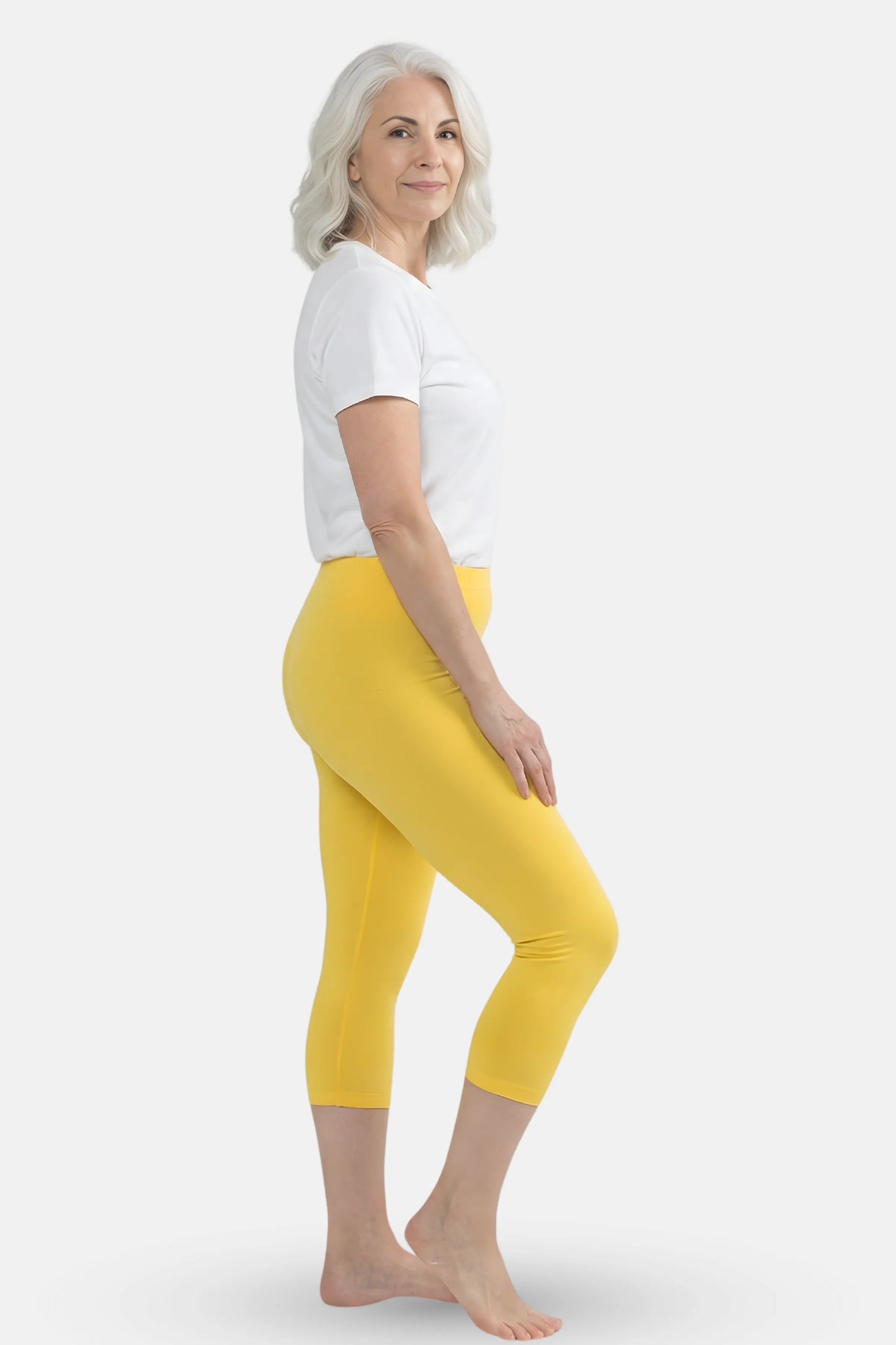 An elder woman wearing bright yellow capri leggings and a white t-shirt against a light gray studio background.