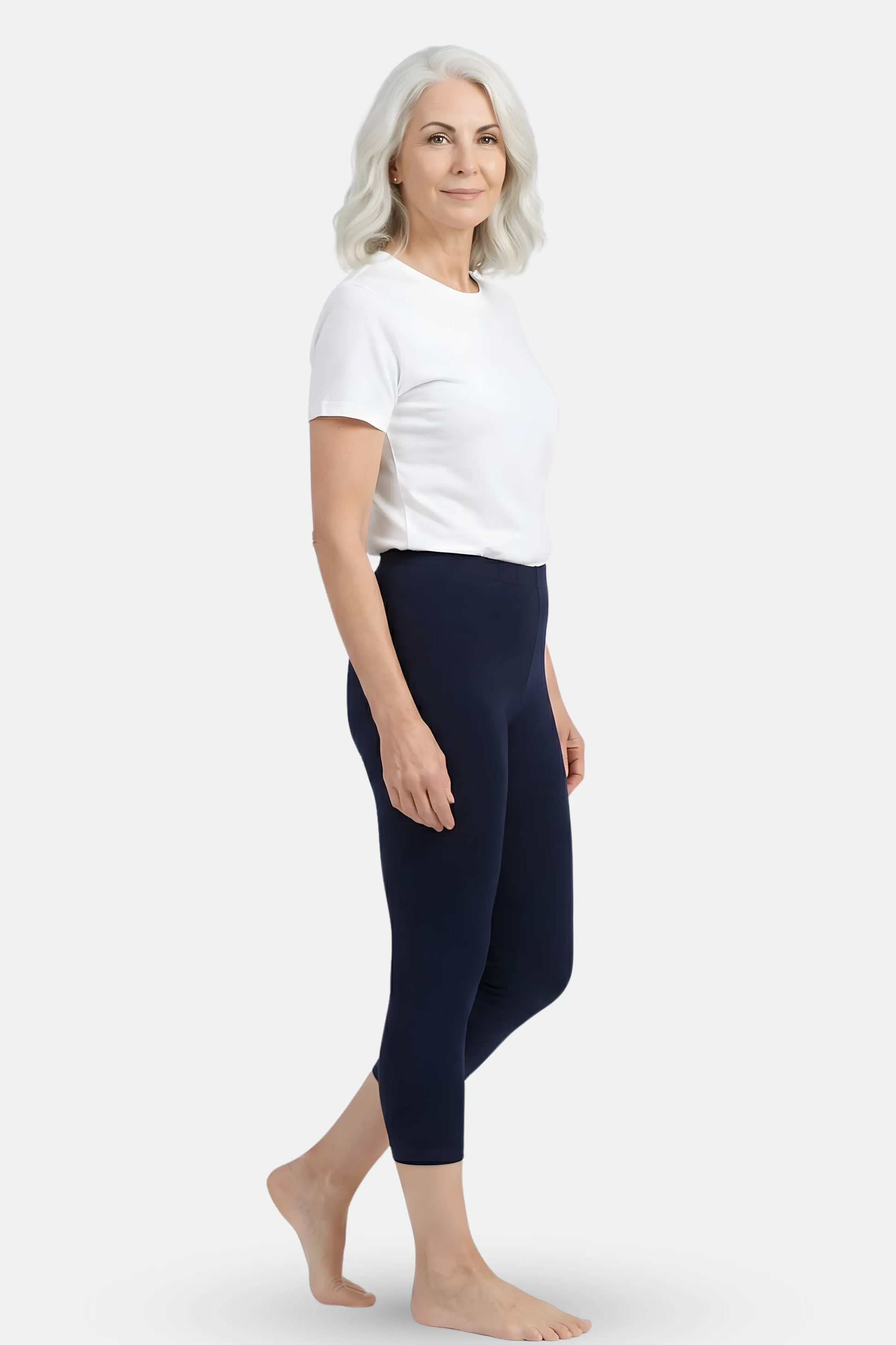 An elder woman wearing a white  t-shirt and dark navy blue high-waisted cropped leggings against a light gray studio background.