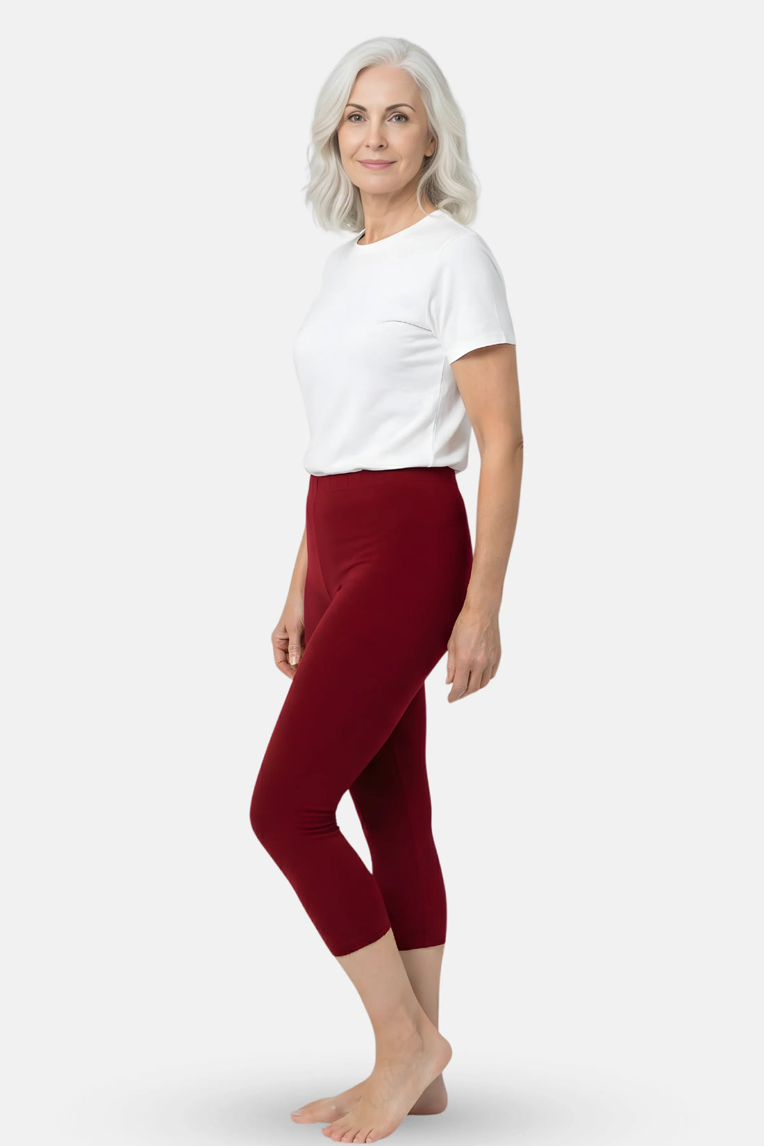 A woman with wearing a white short-top and maroon capri leggings against a light gray studio background.