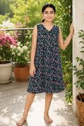 A young girl stands barefoot on a sunlit patio wearing a sleeveless V-neck midi dress with multicolor floral print.