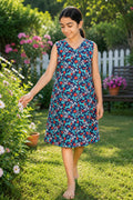 A young girl wearing a sleeveless V-neck dress with a red, blue, and white floral pattern, standing barefoot in a sunny garden with green grass and flowers.