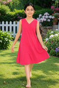 A young girl with dark hair in a ponytail is wearing a sleeveless, V-neck pink dress while walking barefoot on a green lawn in a garden.