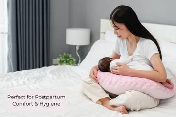 Woman breastfeeding a baby using a pink and white nursing pillow in a bedroom setting.